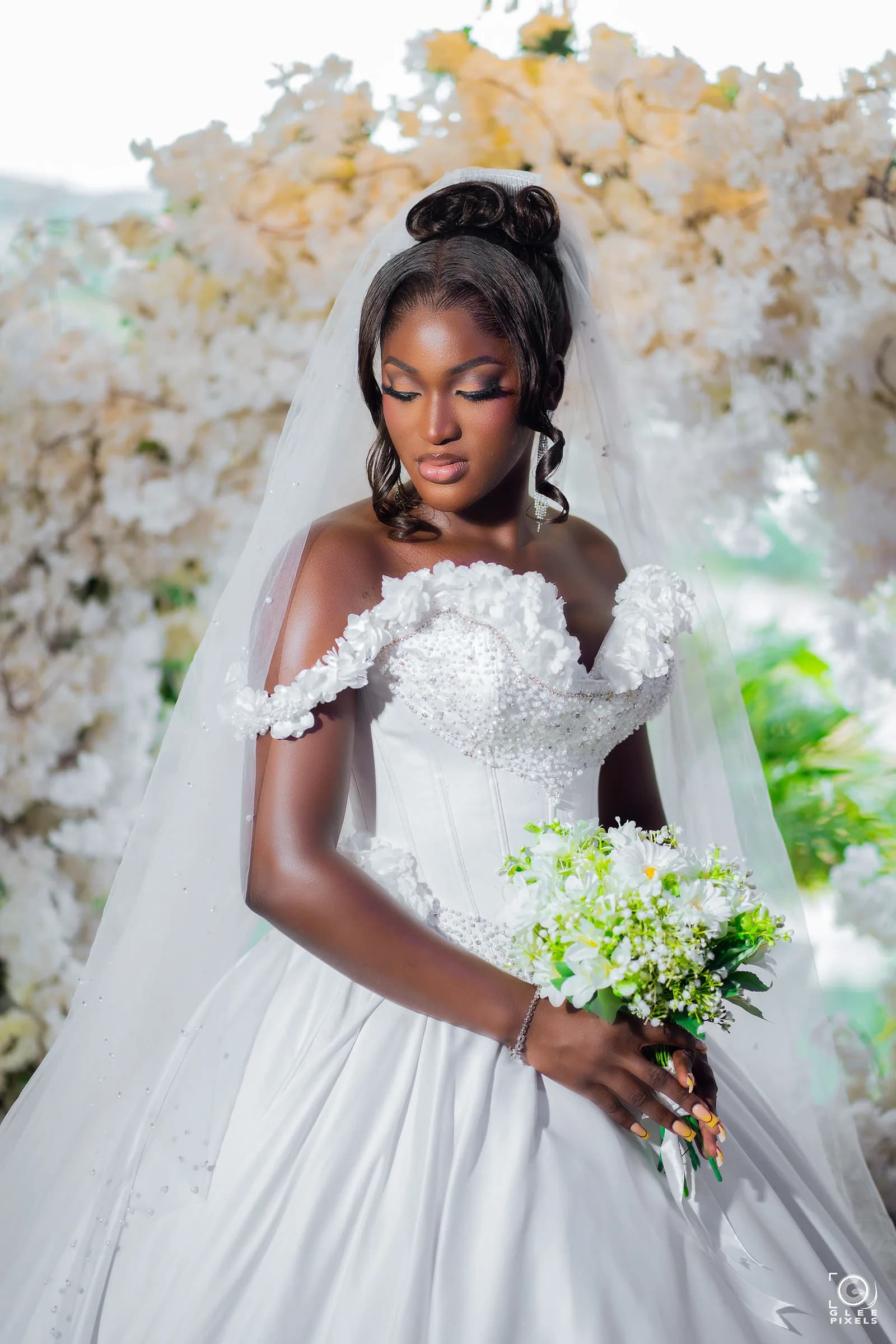 Bride wearing a custom ivory satin ball gown with an off-shoulder bodice appliquéd in three-dimensional floral lace, photographed before an installation of cream and white blossoms.